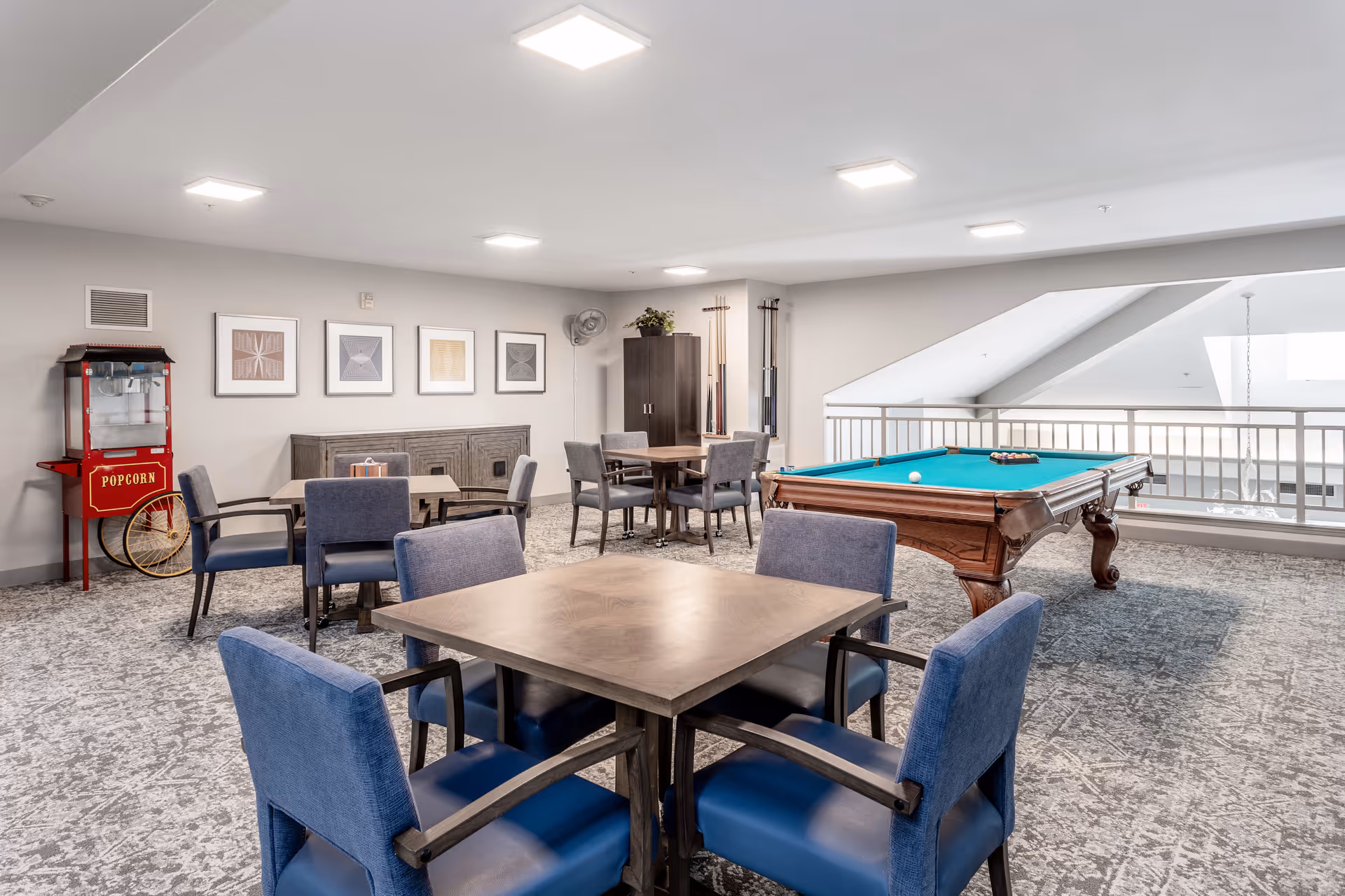 Bright communal game room with a pool table, multiple card tables and blue chairs, and a vintage popcorn machine.