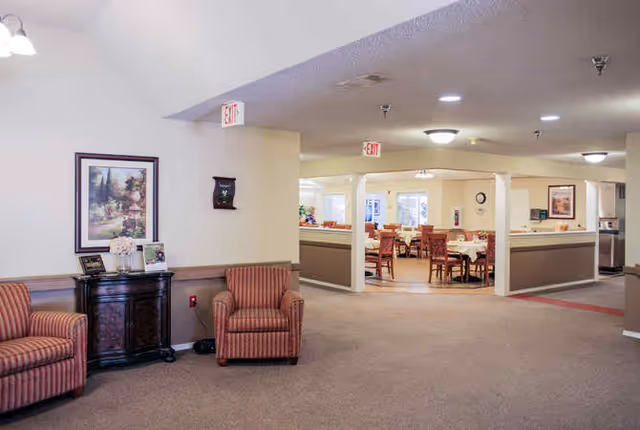 Interior view of a senior living facility showing a seating area with two striped armchairs and a small cabinet with framed pictures and a flower vase. In the background, there is a dining area with tables and chairs, and exit signs are visible on the ceiling.