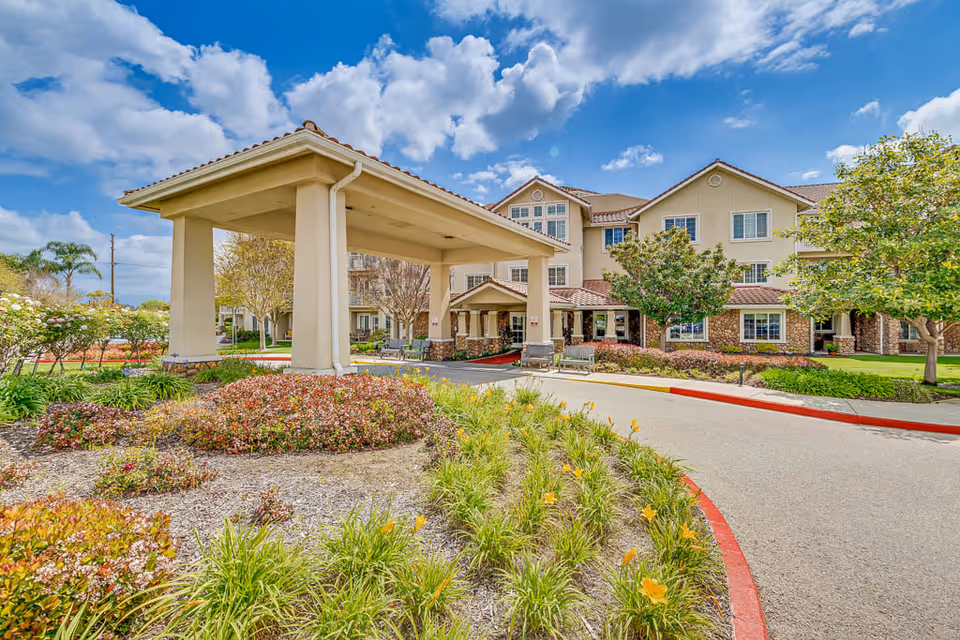 Exterior view of The Palms At Bonaventure Assisted Living & Memory Care building with a covered entrance, surrounded by landscaped gardens and trees under a partly cloudy sky.