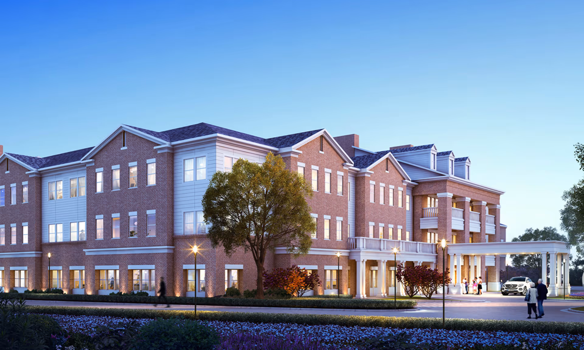 Three-story brick senior living building with a covered entrance, landscaped grounds, and a few people near the driveway at dusk.