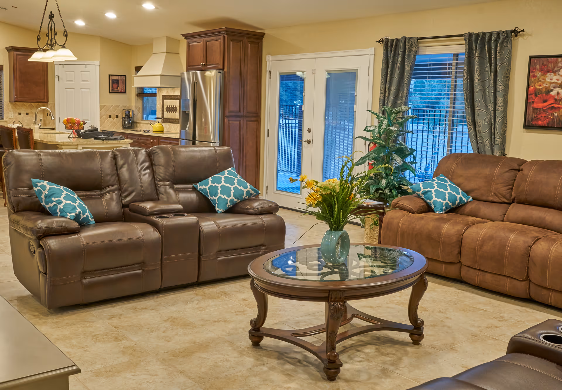 A cozy living room with two brown leather sofas adorned with blue patterned throw pillows, a round glass-top wooden coffee table with a vase of yellow flowers, a potted plant near French doors with blue curtains, and a view into a kitchen area with wooden cabinets and stainless steel appliances.