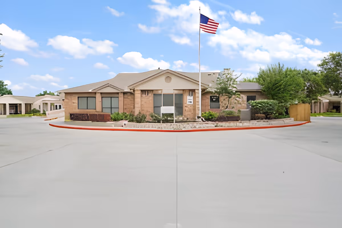 Front exterior of a single-story senior living building with an American flag on a pole and a circular driveway.