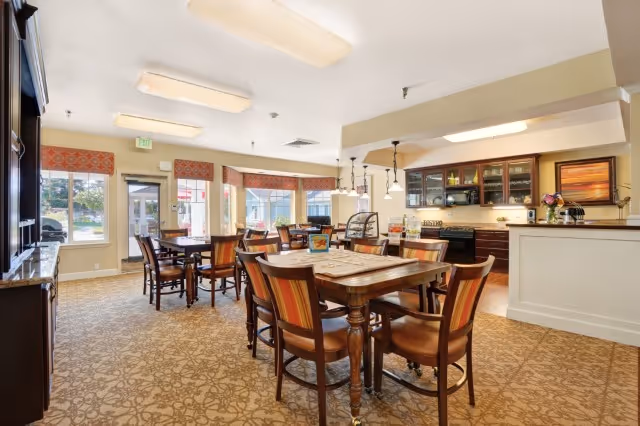 Communal dining area with several wooden tables and chairs, a serving counter/kitchenette, patterned carpet and large windows.