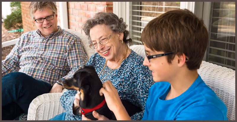 An elderly woman sitting on a white wicker bench on a porch, smiling and holding a small black dog with a red collar, accompanied by a man and a boy who are also smiling and interacting with the dog.