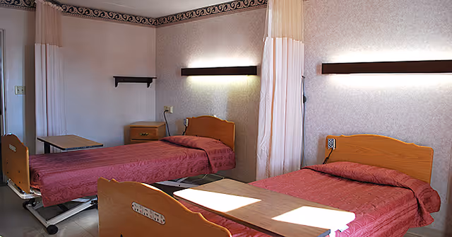 A room with two hospital-style beds with wooden headboards and footboards, each covered with a red bedspread. There are two overbed tables positioned over the beds. The room has light-colored walls with a decorative border near the ceiling, a small wooden nightstand between the beds, and wall-mounted lights above each bed. Privacy curtains hang between the beds.