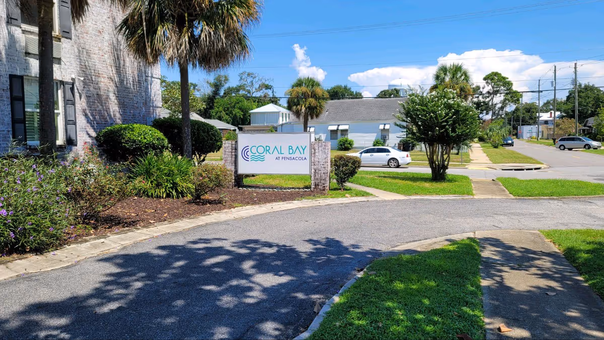 Outdoor view of Coral Bay at Pensacola facility entrance with a sign displaying the facility name, surrounded by palm trees, bushes, and a paved driveway. Residential houses and parked cars are visible in the background under a partly cloudy sky.