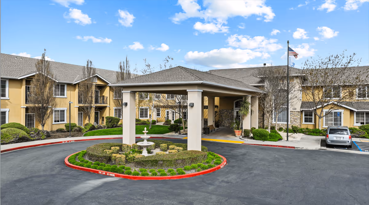 Exterior view of Manteca Assisted Living and Memory Care facility showing a circular driveway with a covered entrance, landscaped greenery, and a two-story building with beige walls and multiple windows under a partly cloudy sky.