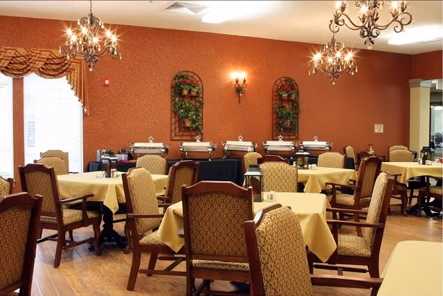 A dining room with several tables covered in yellow tablecloths and surrounded by upholstered wooden chairs. The room has warm red patterned walls with decorative wall sconces and two chandeliers hanging from the ceiling. There are buffet serving trays lined up against the far wall beneath two wall-mounted plant decorations. Large windows with patterned valances allow natural light into the room.