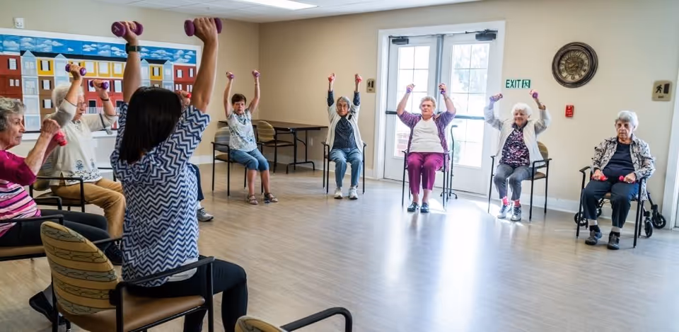 A group of elderly women seated in a circle in a spacious room participating in a seated exercise class, each holding small dumbbells and raising their arms. The room has light-colored walls, a large windowed door, and a colorful mural on one wall.