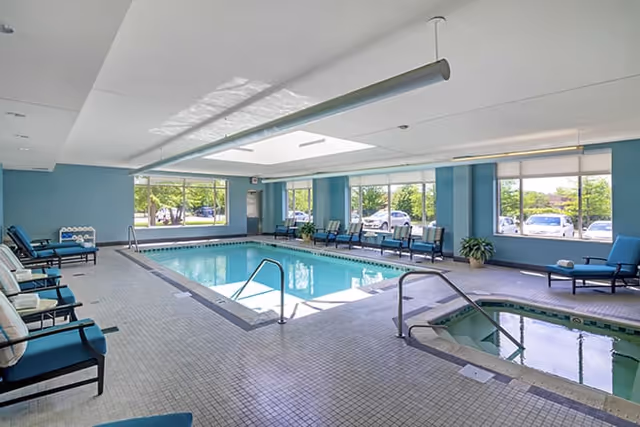 Indoor swimming pool area with large windows letting in natural light. The pool is surrounded by tiled flooring and several cushioned lounge chairs and seating along the walls. There is also a hot tub adjacent to the pool.