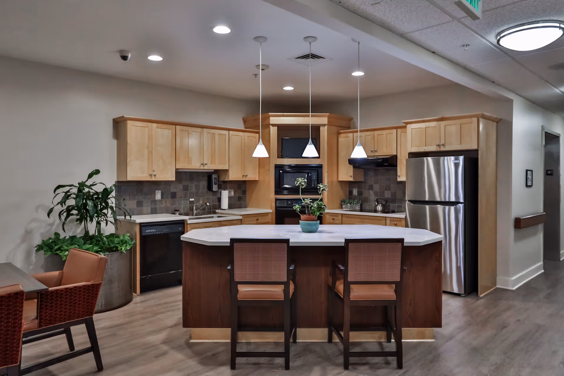 A modern kitchen area in a senior living facility featuring light wood cabinets, a stainless steel refrigerator, a built-in oven and microwave, a dishwasher, and a central island with a white countertop and two chairs. There are three pendant lights hanging above the island and a potted plant on the countertop. The floor is wood, and there is a large potted plant to the left near a seating area with two chairs.