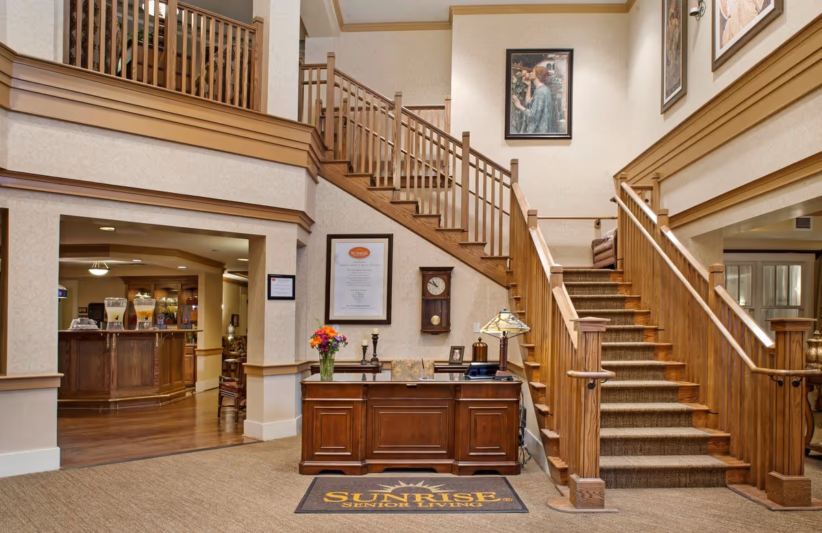 Interior view of a senior living facility lobby with a wooden reception desk, a staircase with wooden railings leading to an upper floor, framed artwork on the walls, and a glimpse into a dining or common area with beverage dispensers.