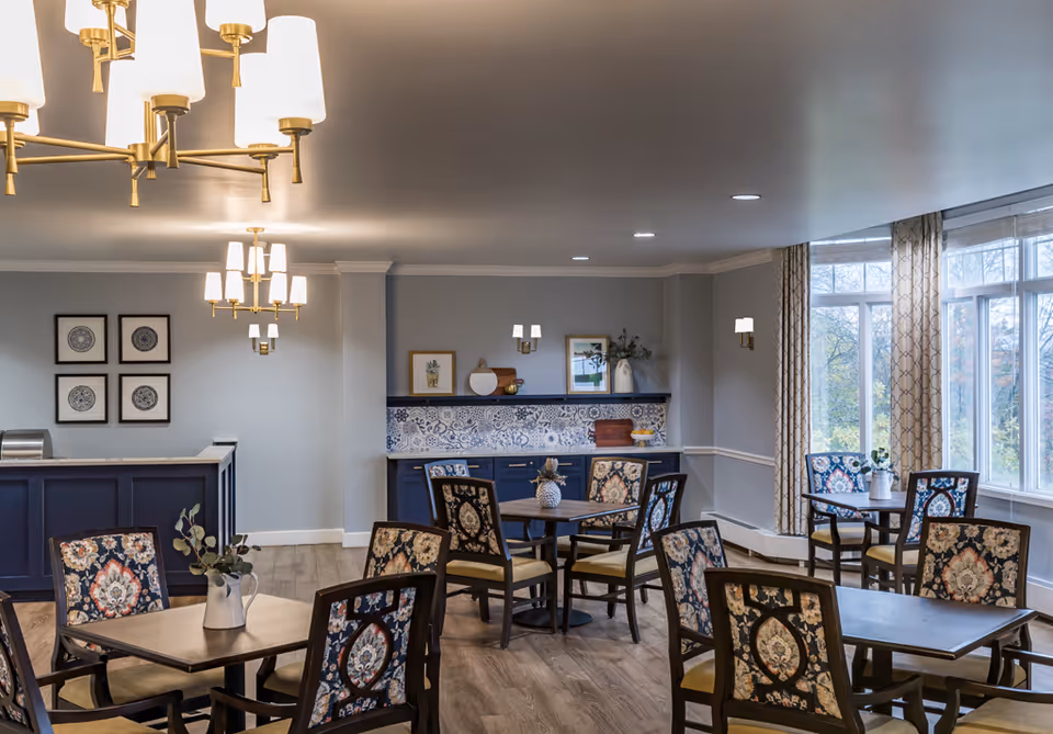 Bright communal dining room with patterned chairs, several tables, chandeliers, and large windows.