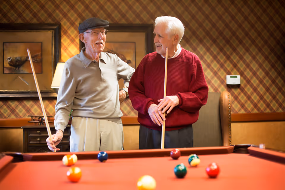 Two elderly men standing by a red pool table in a room with plaid wallpaper and framed artwork. One man is wearing a gray shirt and cap, holding a pool cue, while the other man is wearing a red sweater and holding a pool cue, both smiling and engaged in conversation.