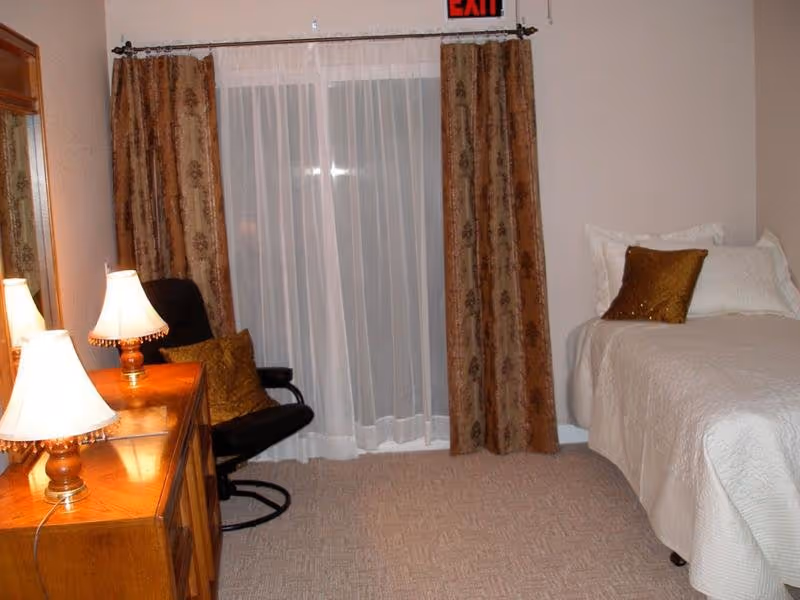 A cozy bedroom with a single bed covered in white bedding and a brown decorative pillow. Next to the bed is a wooden dresser with two lit lamps on top. A black chair with a brown cushion is positioned near a window with sheer white curtains and brown patterned drapes. An exit sign is visible above the window.