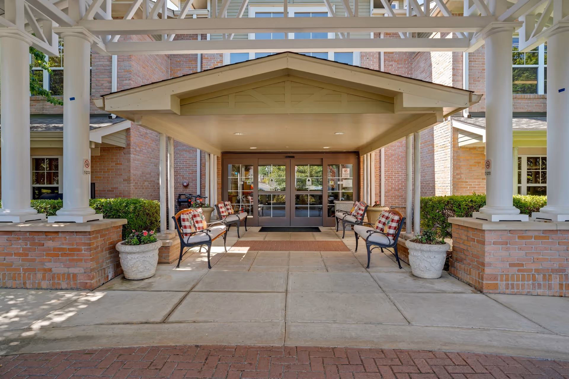 Entrance to a senior living facility with a covered porch supported by white columns. There are benches with plaid cushions on either side of the walkway leading to glass double doors. Brick walls and greenery surround the entrance area.