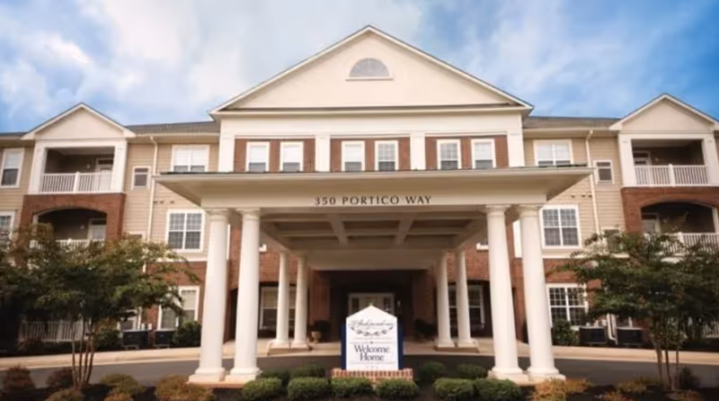 Front exterior view of a three-story senior living facility building with a covered entrance supported by white columns. The address 350 Portico Way is displayed above the entrance. There are trees and shrubs in front of the building and a sign that reads 'Welcome Home'.