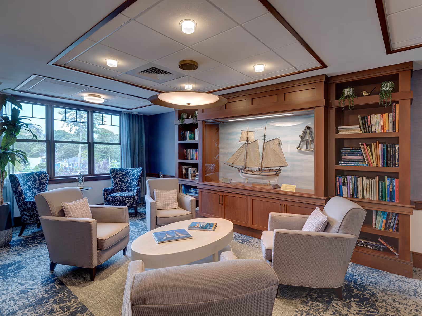 A cozy living room area with four beige armchairs arranged around an oval coffee table with books on it. Behind the chairs is a built-in wooden bookshelf filled with books and decorative plants. A large model sailboat is displayed in a recessed wooden cabinet on the wall. Two patterned blue armchairs are positioned near a window with blue curtains, letting in natural light. The ceiling has recessed lighting and a central round light fixture.
