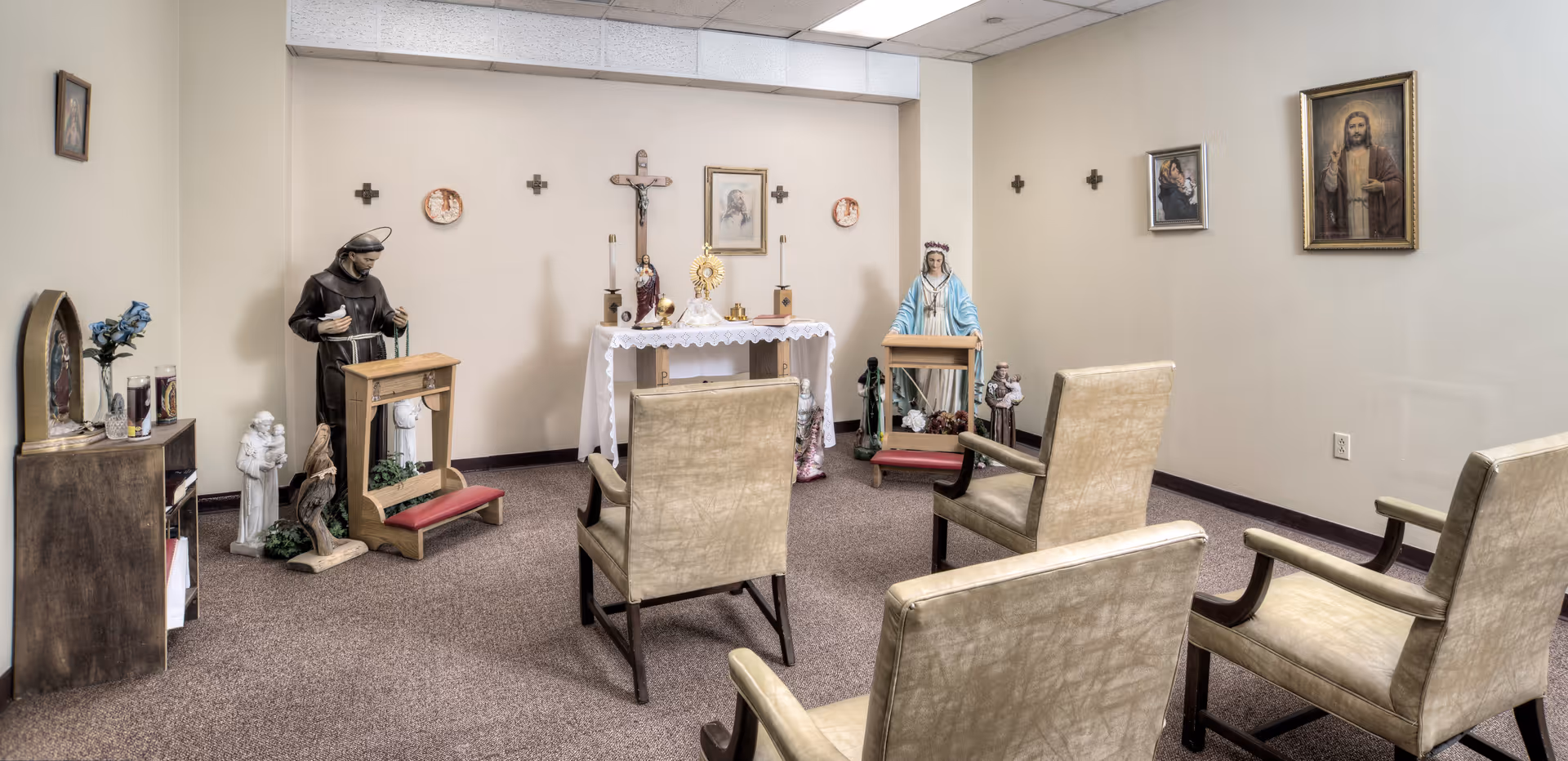 A small prayer room with religious statues, including a crucifix, a statue of the Virgin Mary, and other saint figures. There are four beige chairs arranged facing a small altar covered with a white cloth. The walls are decorated with small crosses and framed religious pictures.