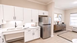Bright and modern kitchen area with white cabinets, a stainless steel refrigerator, a sink, and a countertop. Adjacent to the kitchen is a living space with a sideboard, decorative items, and a window with blinds.