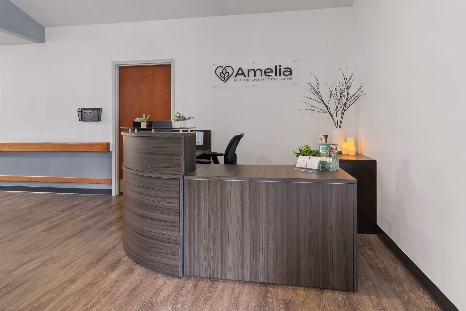 Lobby reception area with a curved wood desk, decorative plants and an 'Amelia' sign on the wall.