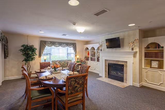 A cozy living room area with a wooden dining table surrounded by six chairs with floral upholstery. In the background, there is a floral-patterned sofa beneath a window with draped curtains. The room features a white fireplace with a mounted flat-screen TV above it, built-in shelves with books and decorative items, and a potted plant in the corner. The carpeted floor and soft lighting create a warm and inviting atmosphere.