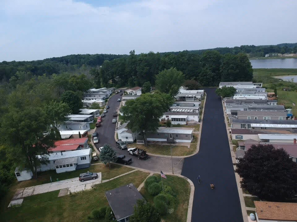 Aerial view of a mobile home community with multiple single-story homes lined along paved roads, surrounded by trees and greenery, with a lake visible in the background under a partly cloudy sky.