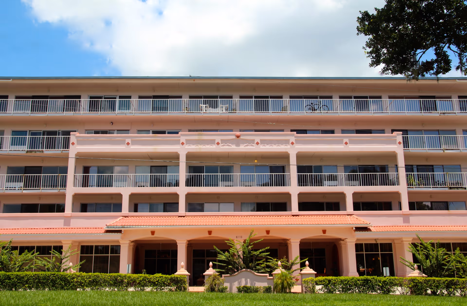 Exterior view of a multi-story senior living facility building with balconies on each floor, pink walls, and a red-tiled roof entrance. There is green grass and shrubs in front of the building under a partly cloudy blue sky.