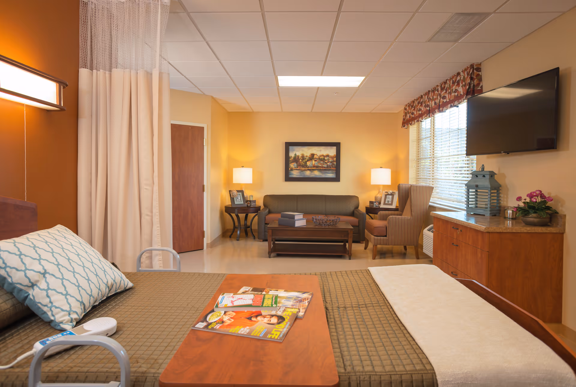 A cozy senior living facility room featuring a bed with a patterned pillow and a wooden overbed table holding magazines. The room has warm beige and orange walls, a sitting area with a sofa, armchair, coffee table, and two lamps. A flat-screen TV is mounted on the wall above a wooden cabinet with decorative items. A window with blinds and a floral valance lets in natural light.