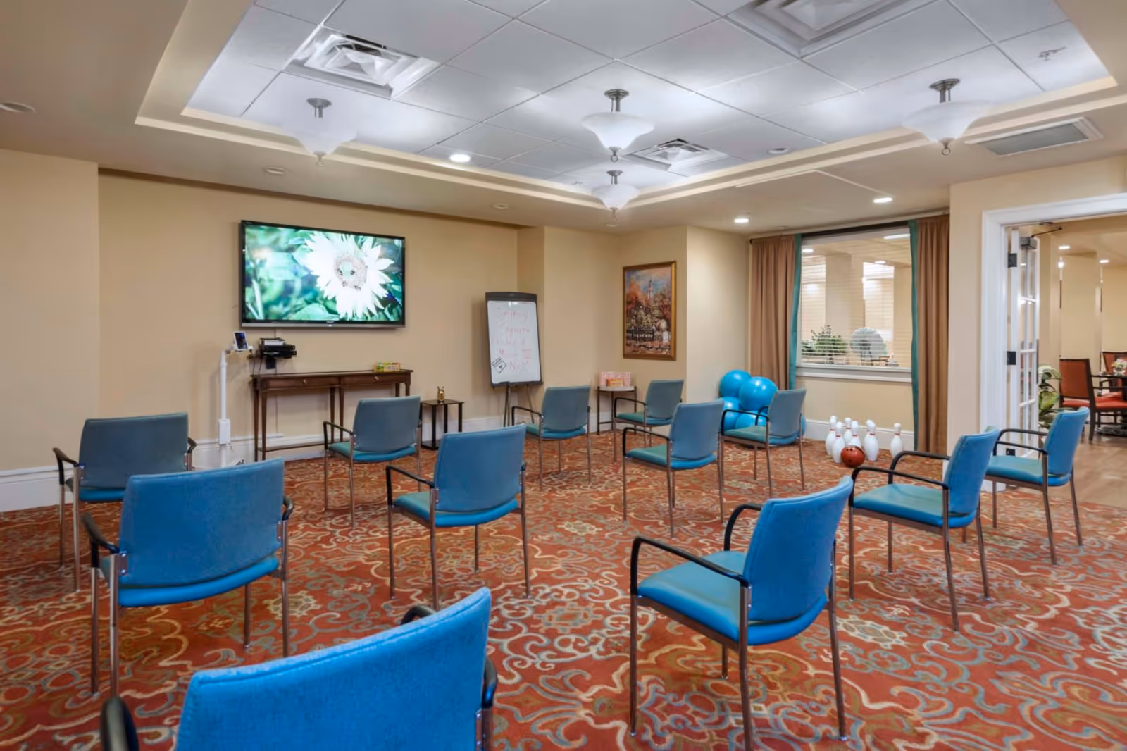A room with blue chairs arranged in a circle on a patterned carpet. A large TV screen on the wall displays a close-up image of a white flower. There is a small table beneath the TV and a whiteboard with writing on it. In the corner, there are several blue exercise balls and a set of bowling pins with a bowling ball. The room has beige walls, a decorative ceiling with lights, and a window with brown curtains looking into another room.