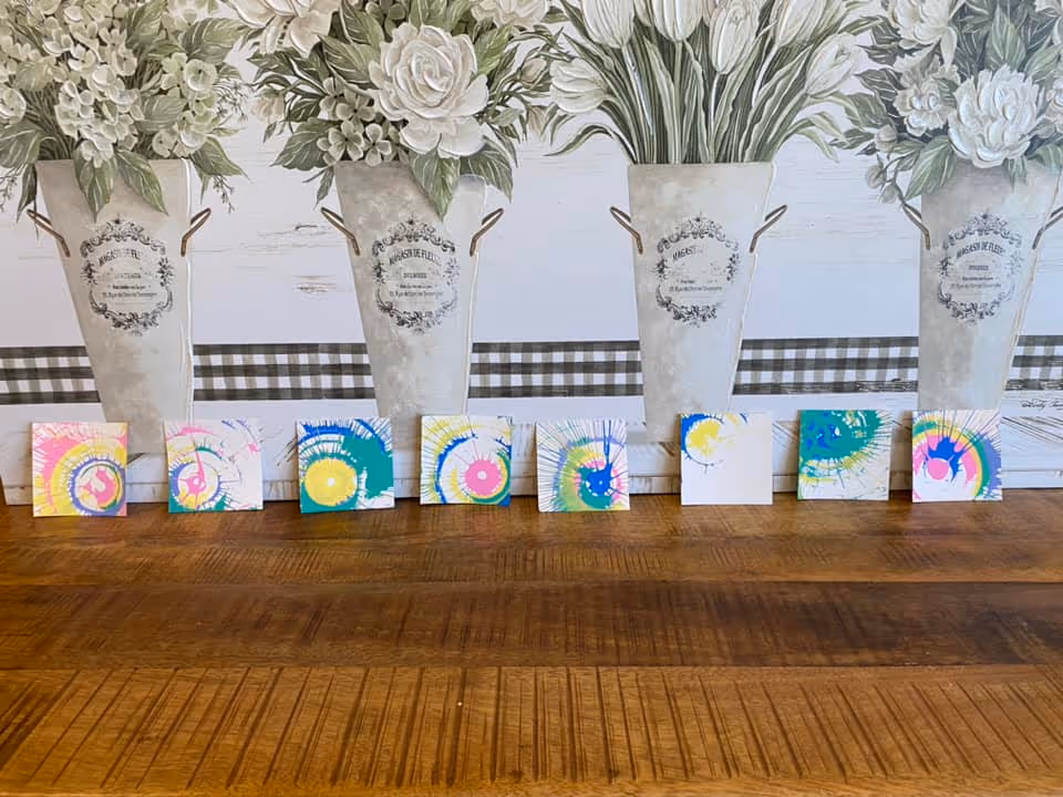 A wooden table with eight small square canvases featuring colorful spiral paint designs arranged in a row. Behind the canvases is a decorative wall with large floral artwork depicting white flowers in tall vases with a checkered pattern strip in the background.