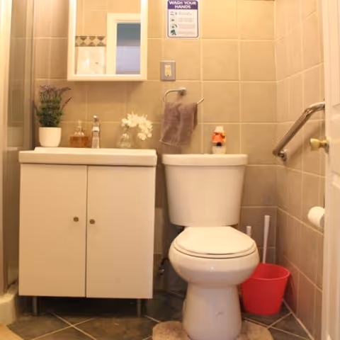 Small bathroom with beige tiled walls featuring a white toilet and a white vanity cabinet with a sink. Above the sink is a mirrored medicine cabinet. There is a towel hanging on a ring next to the toilet, a red bucket on the floor, and a grab bar on the wall beside the toilet. A sign above the towel ring reads 'WASH YOUR HANDS'.
