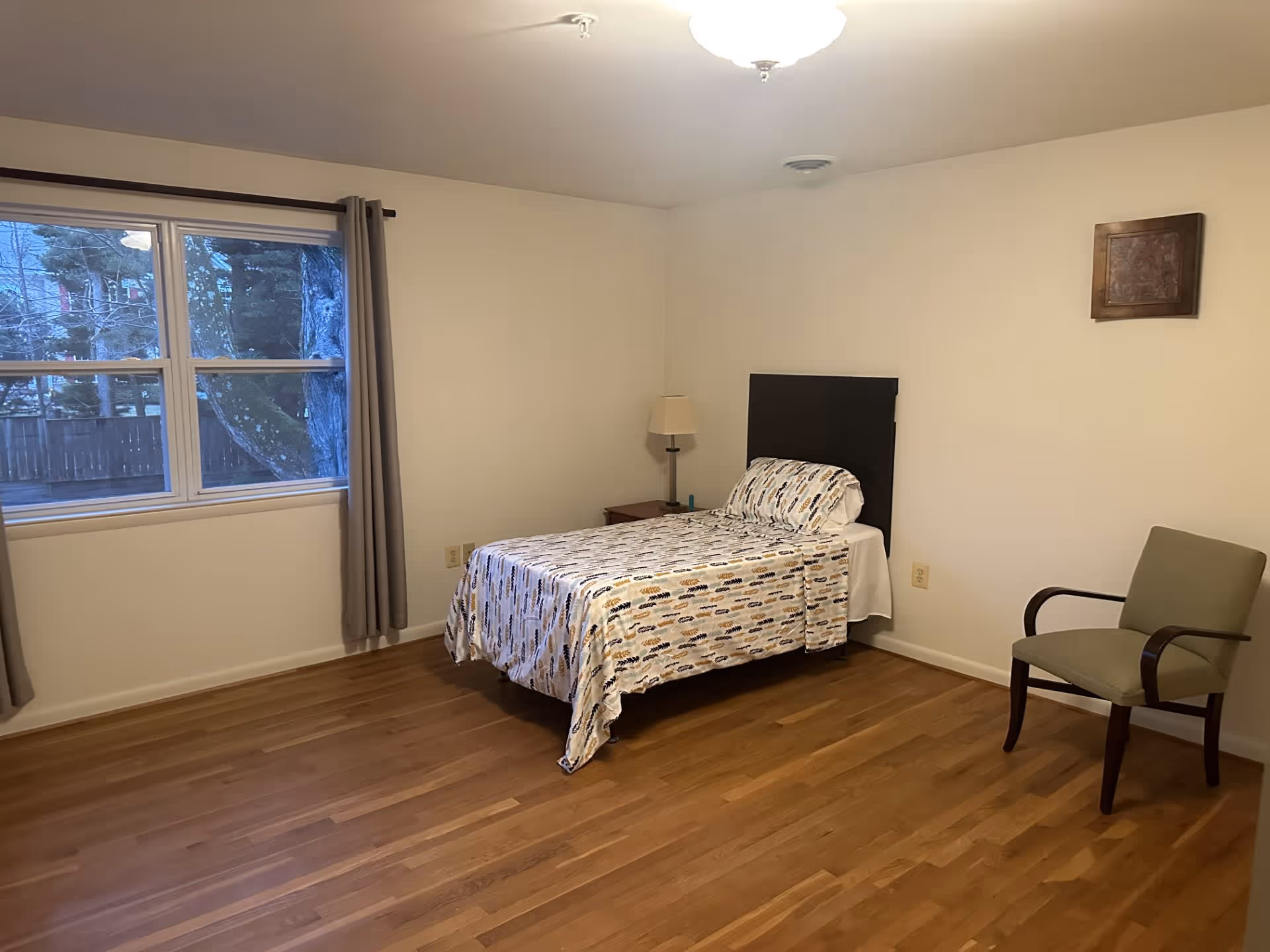 A simple bedroom with a single bed covered with a patterned bedspread, a wooden nightstand with a lamp, a green upholstered chair with wooden arms, a window with gray curtains showing trees outside, and a wooden floor.