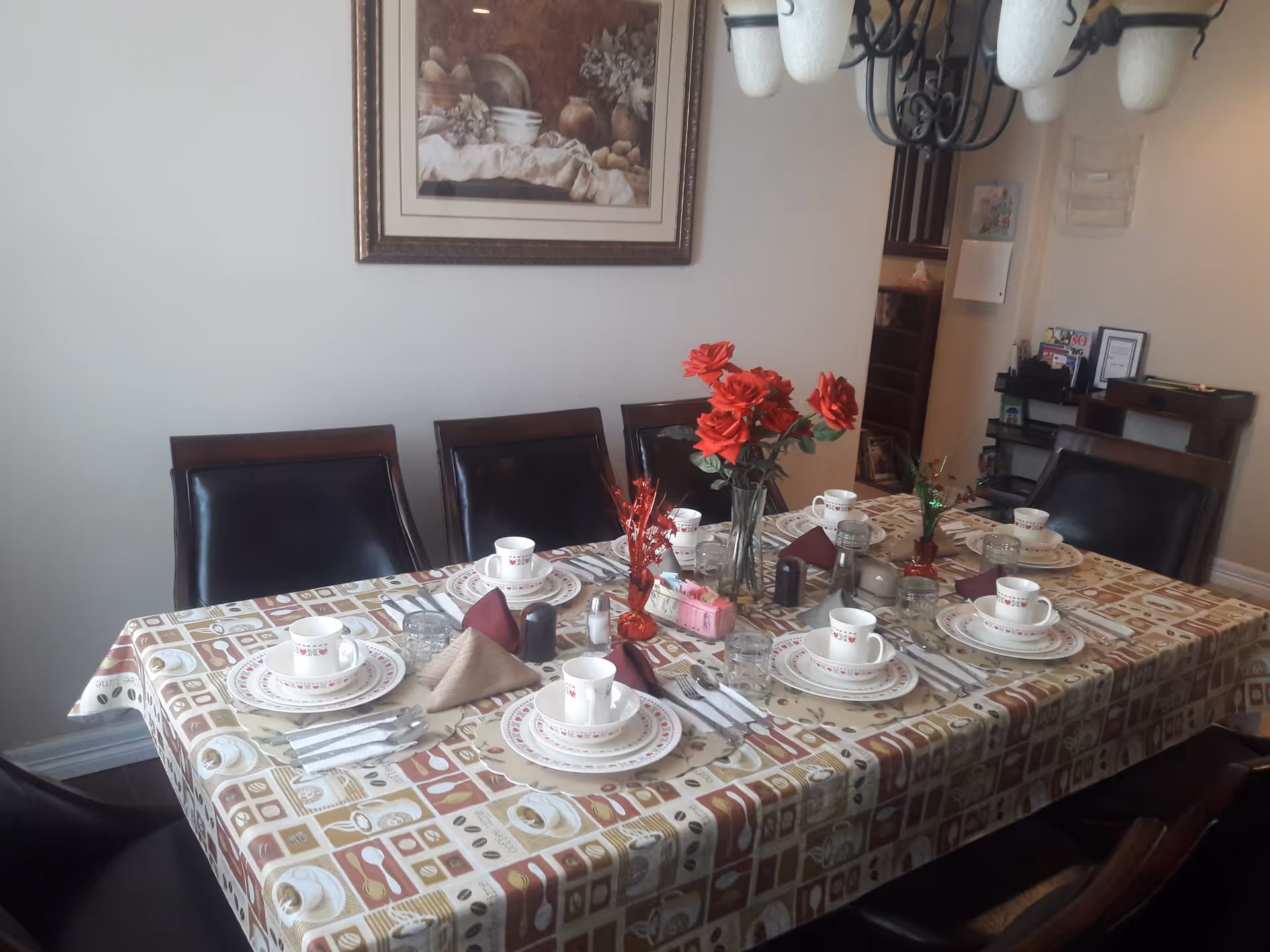 A dining table set for six people with patterned tablecloth, plates, cups, utensils, napkins, and decorative red flowers in vases. The room has dark wooden chairs with black cushions, a framed picture on the wall, and a chandelier overhead.