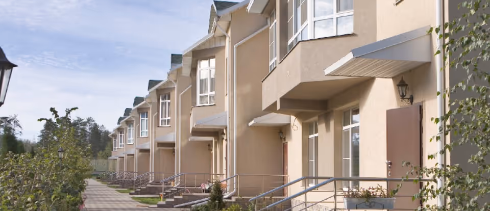Row of beige two-story residential buildings with multiple windows and brown doors, each with a small awning above the entrance. A paved walkway runs alongside the buildings with greenery and trees on the left side under a partly cloudy sky.