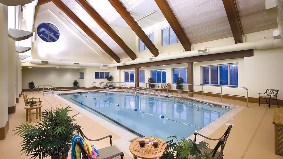 Indoor swimming pool with clear water, surrounded by beige flooring and several chairs and tables with plants. The room has large windows, wooden beams on the ceiling, and a round window near the roof peak.