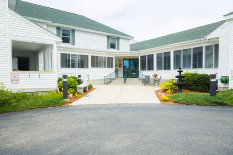 Front entrance of a white two-story nursing home building with a paved walkway, small fountain, and landscaped beds.