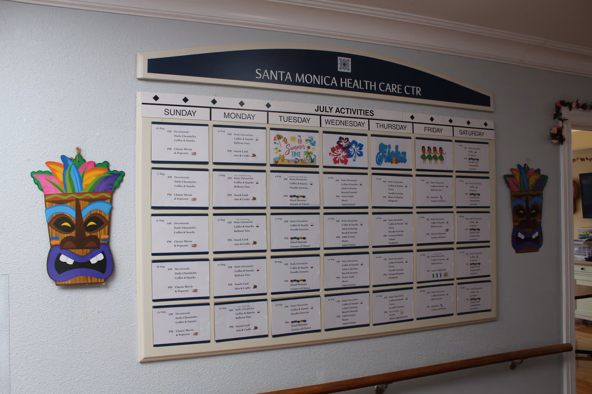 A bulletin board displaying the July activities schedule at Santa Monica Health Care Center. The schedule is organized by day of the week with various daily activities listed. The board is mounted on a white wall with colorful tiki mask decorations on either side. A wooden handrail runs along the wall below the board.