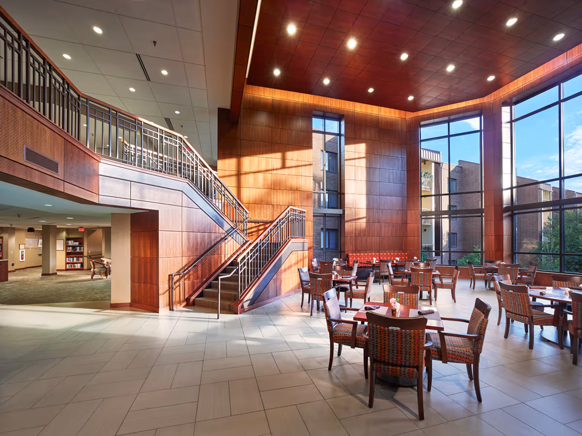 A spacious, well-lit common area with large floor-to-ceiling windows allowing natural light to fill the room. The space features multiple tables and chairs arranged for dining or socializing, with a wooden staircase and wood-paneled walls adding warmth to the modern interior. Outside the windows, parts of a brick building and greenery are visible.