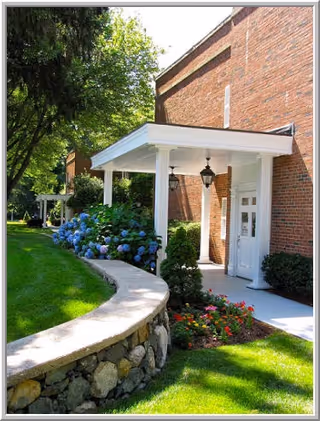 Brick building entrance with a white columned portico, curved stone wall, green lawn, and flowering shrubs.