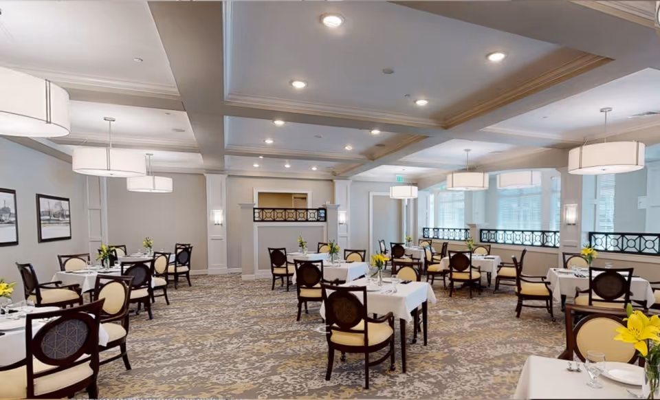 A spacious and elegant dining room with multiple tables covered in white tablecloths, each set with plates, glasses, and yellow flower centerpieces. The room features beige walls, large windows with blinds, decorative ceiling lights, and patterned carpet flooring. Dark wooden chairs with cream-colored cushions surround the tables.