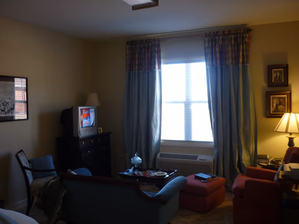 Cozy living room with chairs and ottoman, a TV on a dresser, a window with curtains and an air-conditioning unit beneath it.