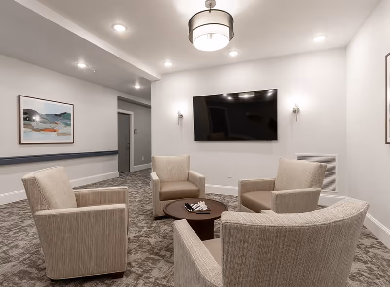 A cozy seating area in a senior living facility with four beige armchairs arranged around a small round table with a chessboard on it. A large flat-screen TV is mounted on the wall, flanked by two wall sconces. The room has neutral-colored walls, carpeted flooring, a framed abstract painting on one wall, and recessed ceiling lights.