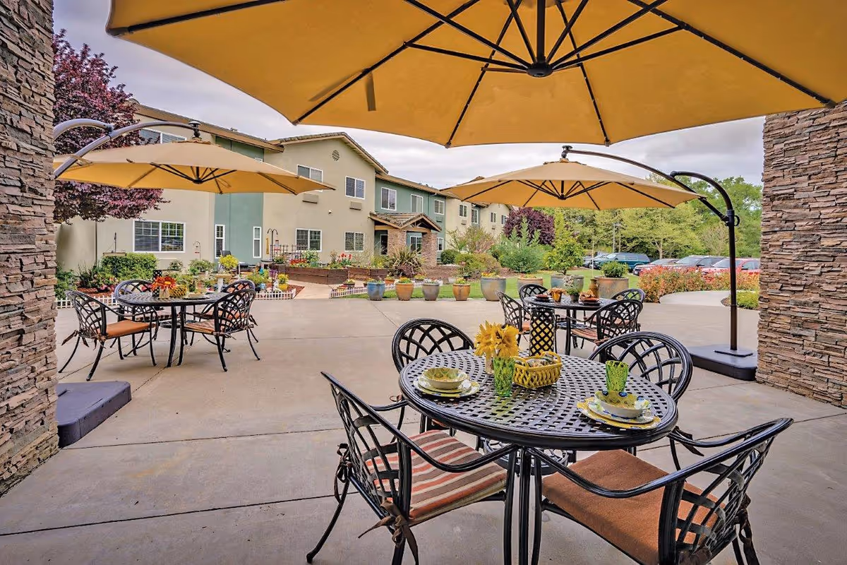 Outdoor patio area at Eskaton Granite Bay with several black metal tables and chairs under large yellow umbrellas. The tables are set with plates, cups, and decorative items. In the background, there are potted plants, a garden area, and a two-story building with green and beige walls. Trees and parked cars are visible further back.