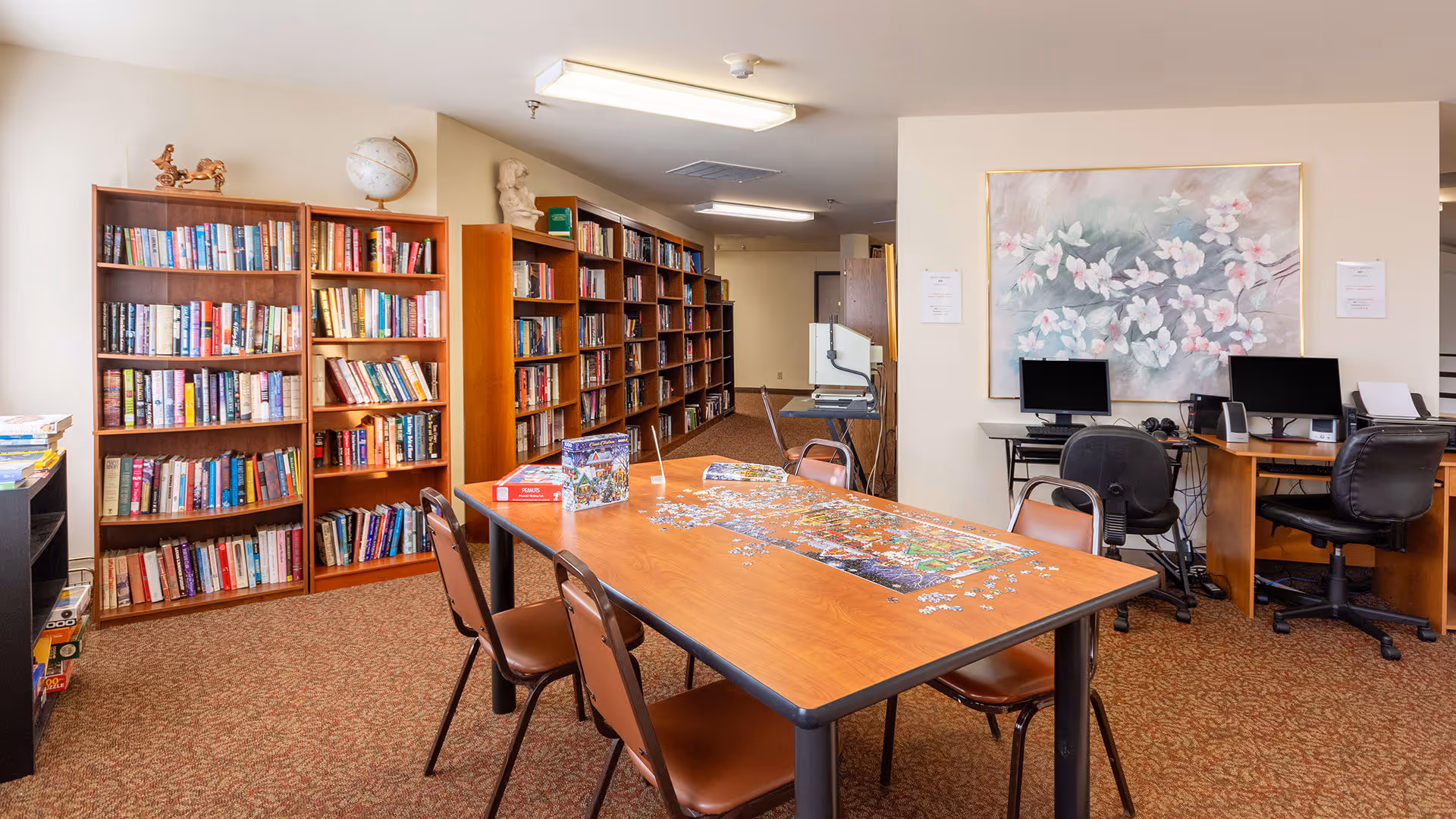 A cozy room with several bookshelves filled with books along the left wall. In the center, there is a rectangular wooden table with a partially completed jigsaw puzzle and four brown chairs around it. On the right side, there are two desks with computer monitors and office chairs. A large floral painting hangs on the wall above the desks. The room has carpeted flooring and fluorescent ceiling lights.
