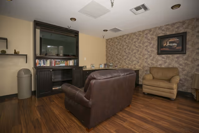 A cozy living room area with a brown leather loveseat facing a large flat-screen TV mounted on a black entertainment center with shelves holding books and decorative items. To the right is a beige armchair against a wall with patterned wallpaper and a framed picture above it. The floor is wooden, and there is a trash can and wall shelves on the left side.