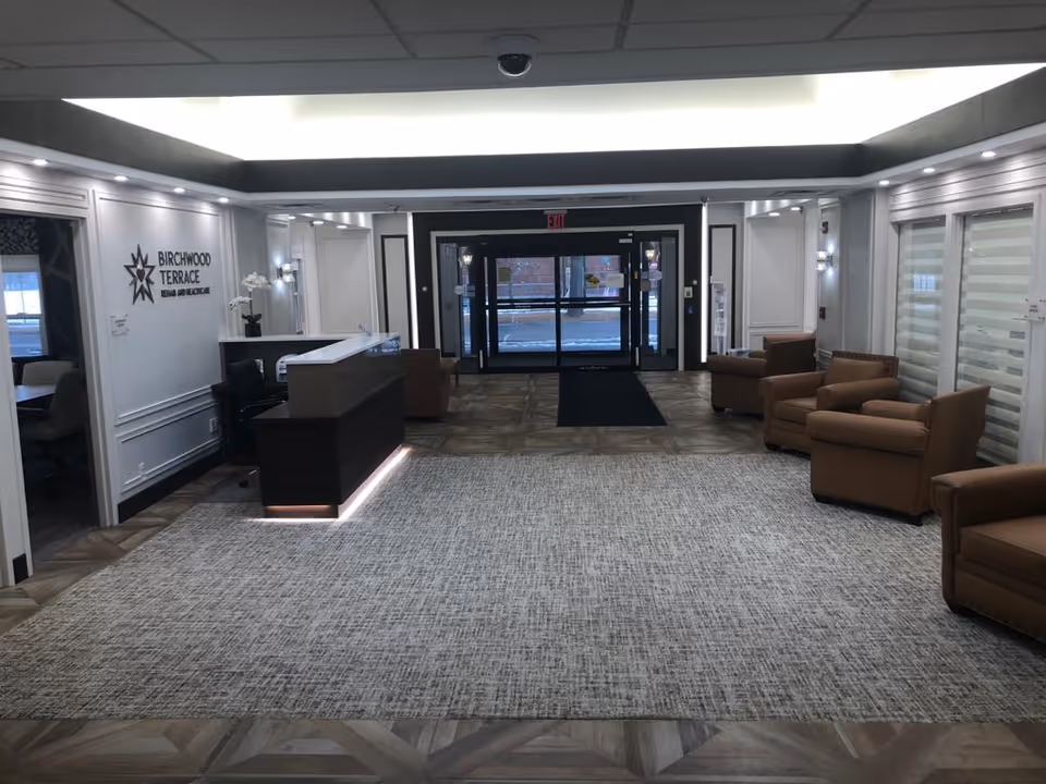 A spacious and well-lit lobby area of Birchwood Terrace Rehab and Healthcare featuring a reception desk on the left side, several brown armchairs arranged along the right wall, a large carpet covering the floor, and glass double doors at the far end leading outside.