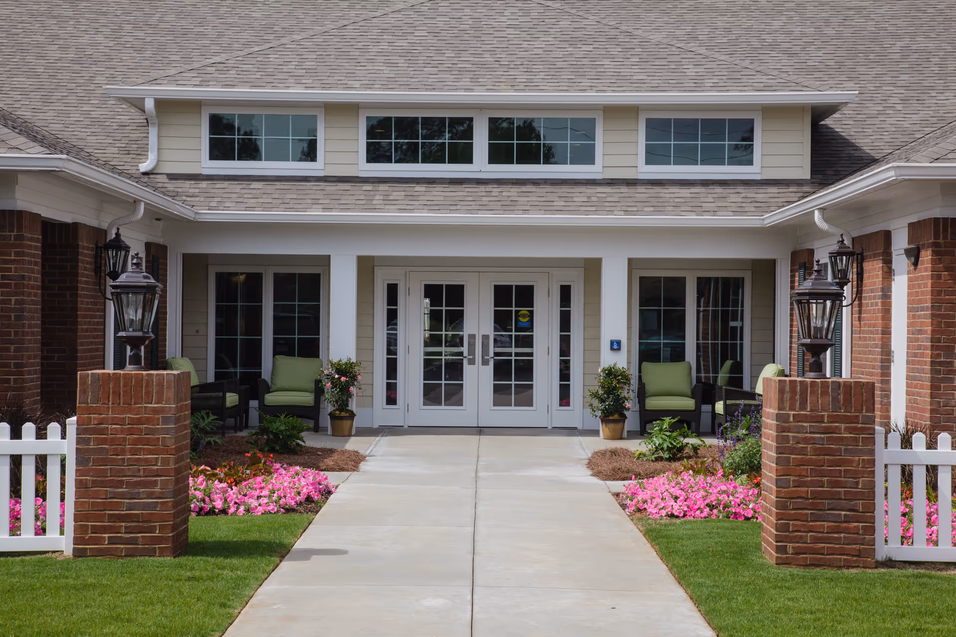 Front entrance of a senior living facility with a concrete walkway leading to double glass doors. The entrance is flanked by brick pillars with lantern-style lights, green cushioned chairs, potted plants, and flower beds with pink flowers. The building has beige siding and a shingled roof with three windows above the entrance.