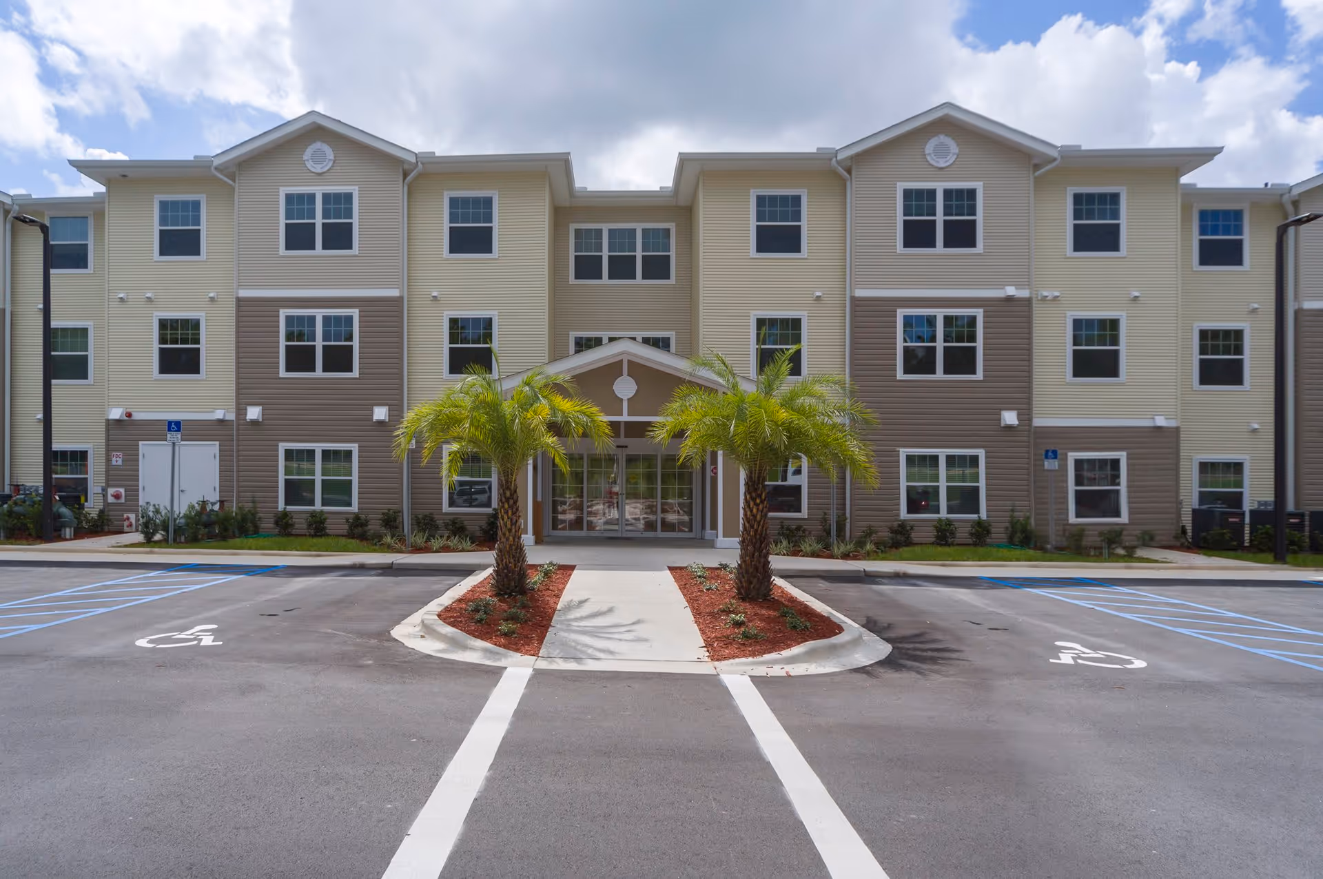 Three-story beige and brown apartment building with a central entrance flanked by two palm trees and marked accessible parking in front.