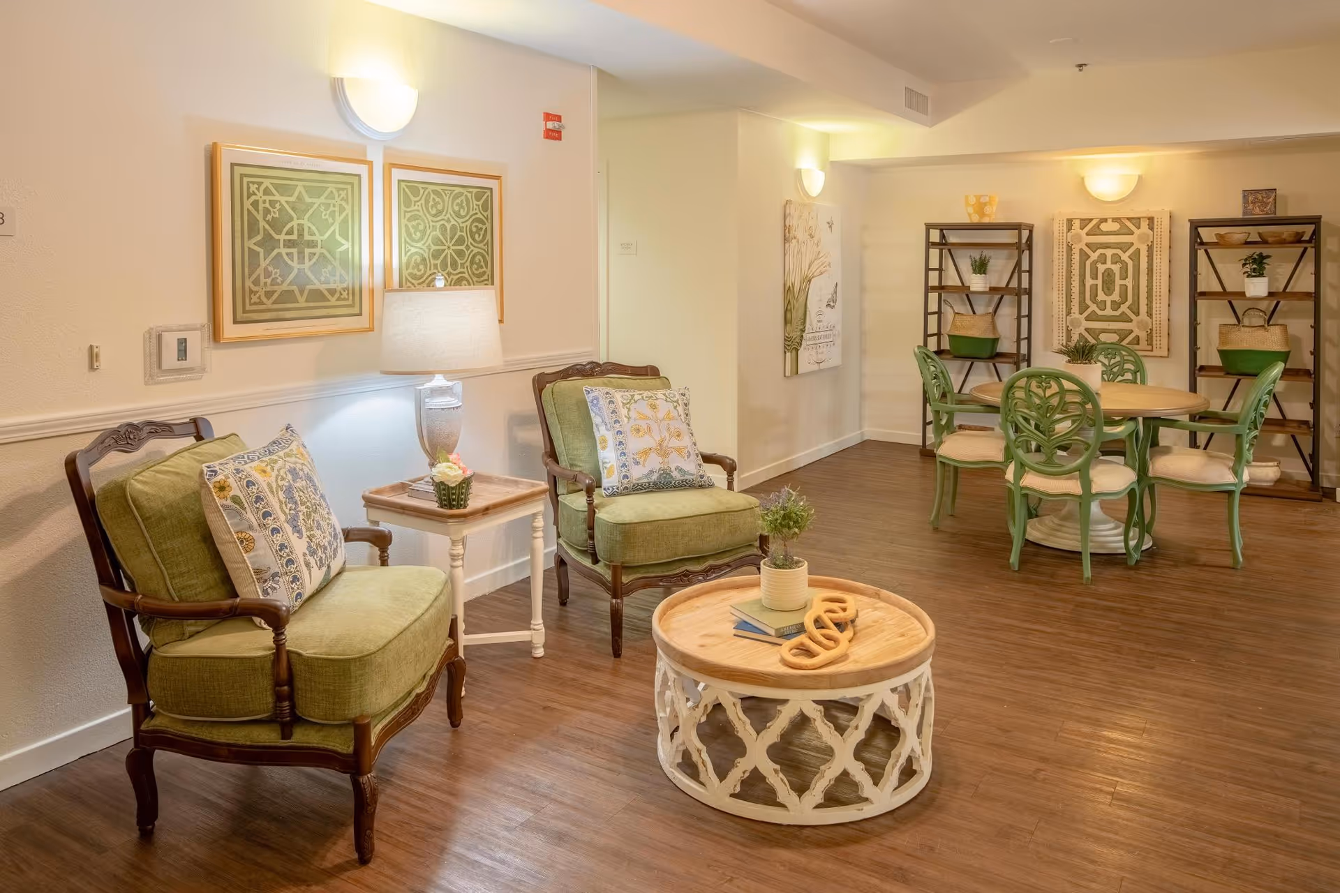 A cozy common area with two green cushioned armchairs with patterned pillows, a small side table with a lamp and flower pot, a round coffee table with decorative items, and a dining table with four green chairs. The room has wooden flooring, light-colored walls, framed artwork, and two shelving units with plants and decorative items.